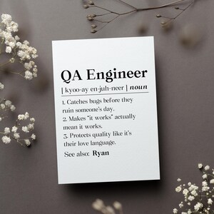 May include: A white card with the text "QA Engineer" in a bold, black font. The card includes a definition, pronunciation, and three statements about the role. The card is surrounded by white flowers and rests on a gray surface.