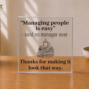 May include: A clear square acrylic desk plaque featuring black printed text that reads: Managing people is easy - said no manager ever - Thanks for making it look that way. Below the text is a line drawing of a coffee mug resting on a stack of papers.