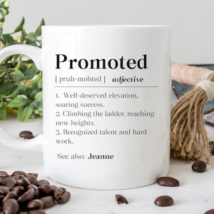 May include: White ceramic mug with a black design that defines the word "Promoted" as an adjective. The definition includes the pronunciation, "[pruh-mohted]", and several bullet points describing the meaning of the word. The mug is sitting on a wooden surface with coffee beans scattered around it.