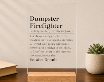Könnte beinhalten: Eine rechteckige Platte aus klarem Acryl auf einem hellen Holzsockel. Die Platte ist mit schwarzem Text bedruckt, der den Begriff Dumpster Firefighter definiert, inklusive Lautschrift, Substantiv-Kennzeichnung und drei nummerierten Punkten.