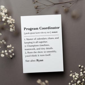 May include: A white card with the definition of "Program Coordinator" in black text. The definition includes the pronunciation, a list of responsibilities, and "See also: Ryan." The card is on a gray surface, with white flowers and sprigs in the corners.
