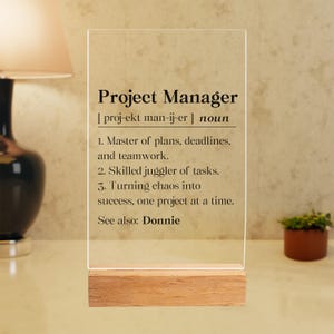 May include: A clear acrylic sign with a wood base that defines "Project Manager" as a noun. The definition includes the phonetic spelling of the word, and lists three responsibilities of a project manager: master of plans, deadlines, and teamwork; skilled juggler of tasks; and turning chaos into success, one project at a time. The sign also includes the text "See also: Donnie".