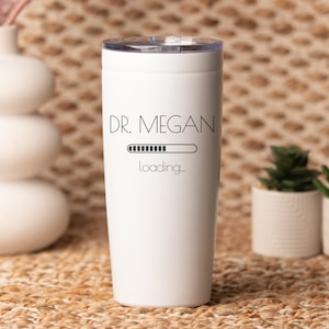 May include: White insulated tumbler with a clear lid. The tumbler features the text "DR. MEGAN" above a loading bar graphic and the word "loading_" in a black, minimalist font. The tumbler is cylindrical and appears to be made of stainless steel.