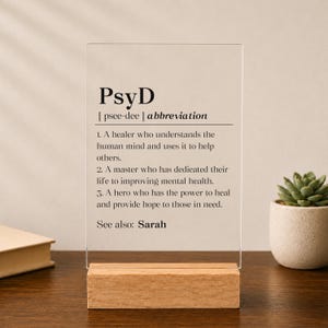 May include: A clear rectangular acrylic sign standing on a light wood base. The sign features black printed text defining PsyD as a healer, master, and hero who improves mental health, with the final line reading: See also: Sarah.