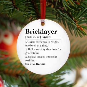 May include: A white, round Christmas ornament with the word "Bricklayer" in black text. The ornament includes a definition of the word, with three descriptive points. A red ribbon is attached for hanging.