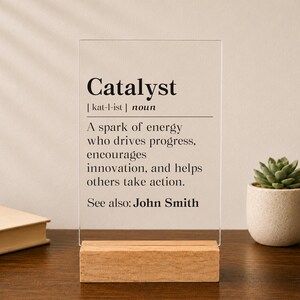 May include: A clear rectangular acrylic plaque standing on a light wood base. The plaque features black printed text defining the word Catalyst as a spark of energy who drives progress, encourages innovation, and helps others take action. See also: John Smith.