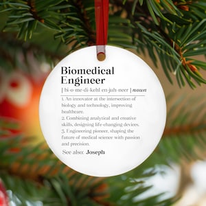 May include: A white, round Christmas ornament with the definition of "Biomedical Engineer" printed in black. The ornament hangs from a red ribbon against a backdrop of green pine needles.