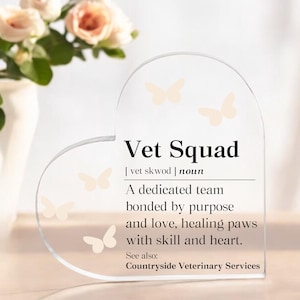 May include: A clear, heart-shaped acrylic plaque with the words "Vet Squad" and a definition. The plaque features light-colored butterfly accents. The definition describes a dedicated veterinary team. A vase of flowers is in the background.