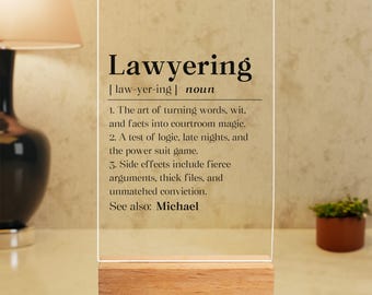 May include: A clear acrylic sign with a wooden base defines "Lawyering" as a noun, describing it as "The art of turning words, wit, and facts into courtroom magic." The sign also lists other aspects of "Lawyering" and ends with "See also: Michael."