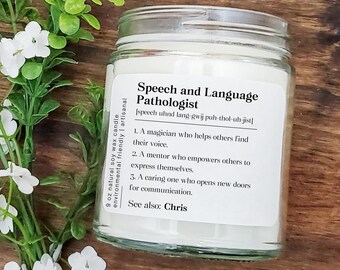 May include: A white candle in a glass jar with a label that reads "Speech and Language Pathologist" and a phonetic pronunciation of the term. The label also includes a description of the profession, stating that a speech and language pathologist is a "magician who helps others find their voice", a "mentor who empowers others to express themselves", and a "caring one who opens new doors for communication". The label also includes the text "See also: Chris".