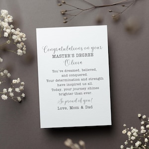 May include: A white card with black text congratulating someone named Olivia on their Master's Degree. The card reads "Congratulations on your MASTER'S DEGREE Olivia You've dreamed, believed, and conquered. Your determination and strength have inspired us all. Today, your journey shines brighter than ever. So proud of you! Love, Mom & Dad"