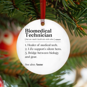May include: A white, round Christmas ornament with the text "Biomedical Technician" in black. The ornament includes a definition and is hanging from a red ribbon against a backdrop of green pine needles.