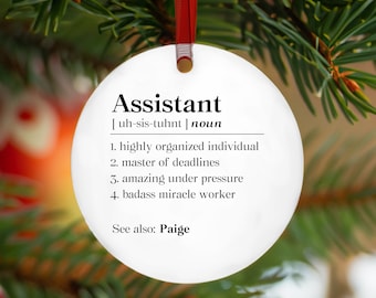 May include: A white, round Christmas ornament with the definition of "Assistant" printed in black. The definition includes qualities like "highly organized" and "badass miracle worker." A red ribbon is attached for hanging. The ornament is displayed on a Christmas tree.