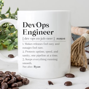 May include: White ceramic mug with the text "DevOps Engineer" and its definition. The definition includes three points about the role. Coffee beans are scattered around the mug.