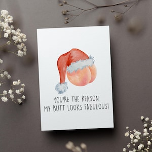 May include: A white greeting card with a watercolor illustration of a Santa hat and peach-shaped bottom. The text reads, "YOU'RE THE REASON MY BUTT LOOKS FABULOUS!" The card is on a gray surface with white flowers.