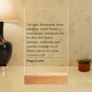 May include: A rectangular clear acrylic plaque standing on a light-colored wooden base. The plaque features black printed text in Spanish: "Así que, hermanos míos amados, estad firmes y constantes, creciendo en la obra del Señor siempre, sabiendo que vuestro trabajo en el Señor no es en vano. 1 Corintios 15:58 Hugo Carlos."