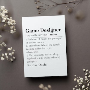 May include: A white card with the definition of "Game Designer" printed in black. The definition includes the pronunciation, a noun, and three descriptive points. The card is on a gray surface, with white flowers and a sprig of a plant in the background.