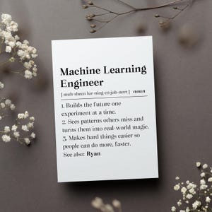 May include: A white card with the definition of a Machine Learning Engineer. The definition includes the phonetic pronunciation, and three descriptive statements. The card is on a gray surface, with white flowers and twigs.