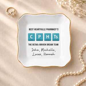 May include: A square white ceramic trinket dish with a gold-trimmed scalloped edge. The center features the text BEST HEARTVILLE PHARMACY'S, four blue periodic table squares spelling C P H Ts, THE DETAIL-DRIVEN DREAM TEAM, and four names.