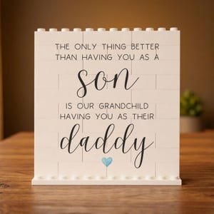 May include: A white brick-style sign with black script that reads "THE ONLY THING BETTER THAN HAVING YOU AS A son IS OUR GRANDCHILD HAVING YOU AS THEIR daddy." A small blue heart is below the word "daddy."
