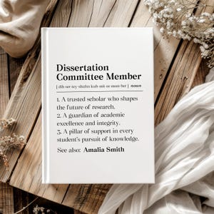 May include: A white book with the text "Dissertation Committee Member" and its definition. The book is on a wooden surface with white fabric and flowers. The definition includes the role of a trusted scholar, guardian of academic excellence, and pillar of support.