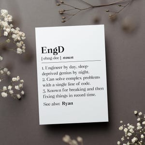 May include: A white card with the definition of "EngD" printed in black. The definition includes the pronunciation, a noun, and three descriptive points about an engineer. The card is surrounded by white flowers and a gray background.