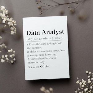 May include: A white card with the definition of "Data Analyst" in black text. The definition includes the pronunciation, a list of responsibilities, and the name "Olivia." The card is on a gray surface, decorated with white flowers and sprigs.