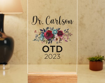 May include: A clear acrylic plaque with a wooden base, featuring a floral design and the text "Dr. Carlson OTD 2023".