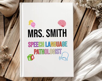 May include: A white notebook with the text "MRS. SMITH SPEECH LANGUAGE PATHOLOGIST" in various colors. The cover also features colorful speech bubbles, a brain, a die, and a speaking outline. The notebook is on a wooden surface.