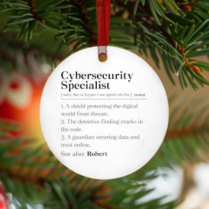 May include: A white, round Christmas ornament with the text "Cybersecurity Specialist" in black. The ornament includes a definition of the term and is hanging from a red ribbon against a backdrop of green pine needles.
