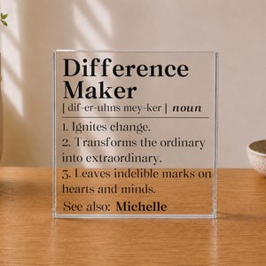 May include: A clear acrylic block sign featuring black printed text that defines the term Difference Maker as one who ignites change, transforms the ordinary into extraordinary, and leaves indelible marks on hearts and minds, with See also: Michelle.