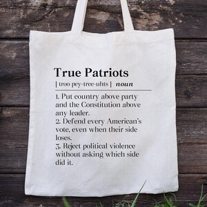 May include: A cream-colored canvas tote bag featuring black printed text that defines the term True Patriots. The text includes a phonetic pronunciation, the noun classification, and three numbered points regarding country, voting, and political violence.