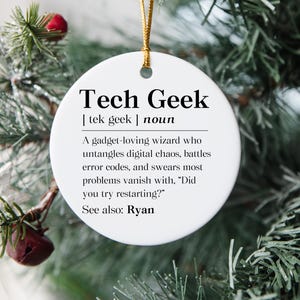 May include: A white, round ceramic ornament with the text "Tech Geek" in black. The ornament includes a definition of "Tech Geek" and is hanging from a gold string, surrounded by Christmas tree branches.