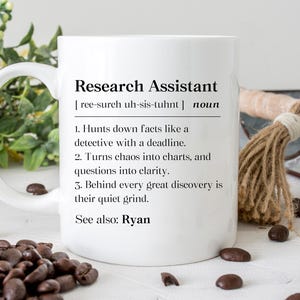May include: White ceramic mug with the text "Research Assistant" and a definition. The definition includes three points about the role. The mug is surrounded by coffee beans and decorative items.