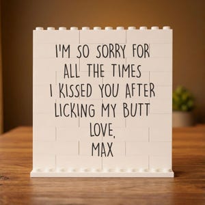 May include: A white brick-style sign with black text that reads "I'M SO SORRY FOR ALL THE TIMES I KISSED YOU AFTER LICKING MY BUTT LOVE, MAX". The sign is on a wooden surface.