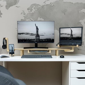 May include: A white desk with a wooden monitor stand holding a laptop and a desktop monitor. The desk has a white drawer unit on the right side and a gray chair in front of it. The wall behind the desk has a gray watercolor world map.