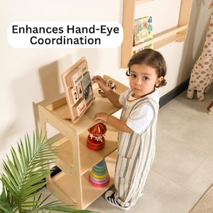 May include: A wooden activity table with a calendar, a red carousel, and a colorful stacking toy. The text "Enhances Hand-Eye Coordination" is displayed above the table. A child in a striped jumpsuit is interacting with the toys.
