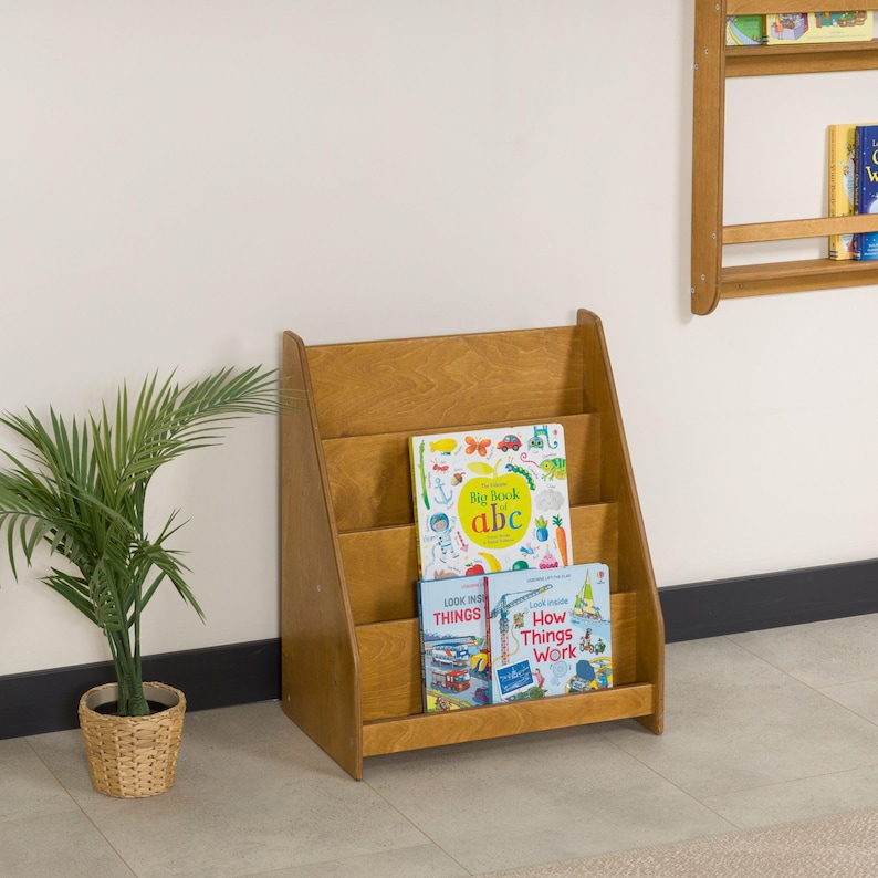 May include: A wooden bookshelf with three shelves, holding children's books. The books include titles like "Big Book abc", "Look Inside Things", and "How Things Work". The bookshelf is made of light-colored wood and is positioned against a white wall.