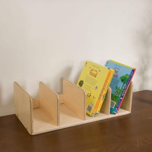 Op de afbeelding: Een lichtgekleurde houten boekenplank met drie compartimenten. Twee boeken zijn zichtbaar, een met een gele kaft met het woord "Food" en een andere met een blauwe kaft met "Our World".