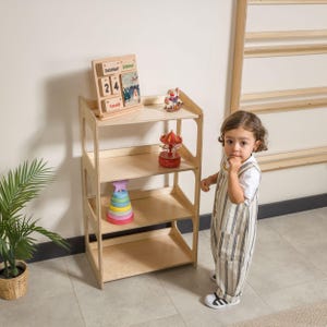 May include: A light-colored wooden children's bookshelf with three shelves. The top shelf holds a wooden calendar, and a small figurine. The middle shelf has a carousel toy. The bottom shelf has a stack of colorful rings. A child is standing next to the bookshelf.