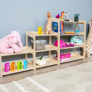 May include: A multi-tiered, light-wood children's bookshelf filled with toys and books. A large pink elephant plush sits on the lowest shelf. Other items include a giraffe, roller skates, and a pink helmet.