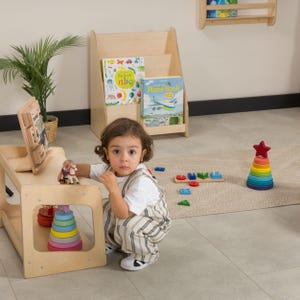 May include: A child's play area featuring a wooden table with a colorful stacking toy and a small figurine. A wooden bookshelf holds children's books. A multi-colored stacking ring toy with a red star on top is also visible.