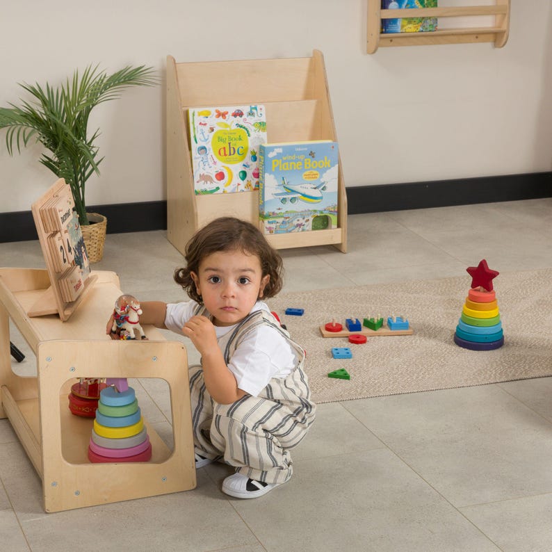 May include: A child's play area featuring a wooden table with a colorful stacking toy and a small figurine. A wooden bookshelf holds children's books. A multi-colored stacking ring toy with a red star on top is also visible.