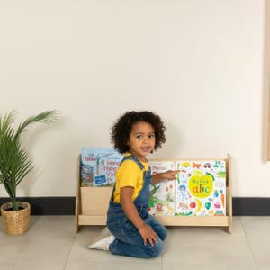 May include: A light-colored wooden bookshelf holding three children's books. A young child in a yellow shirt and denim overalls kneels in front of the bookshelf, pointing at one of the books. A potted plant is to the left.