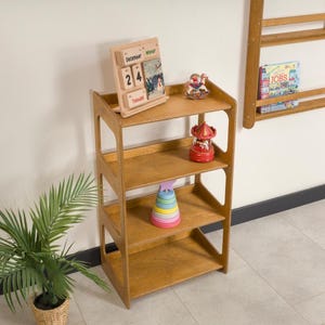 May include: A wooden, four-tiered shelf unit with open sides. The shelves display a wooden calendar, a red carousel toy, and a stack of colorful rings. A small potted plant is in the foreground. A wall-mounted shelf with books is visible in the background.