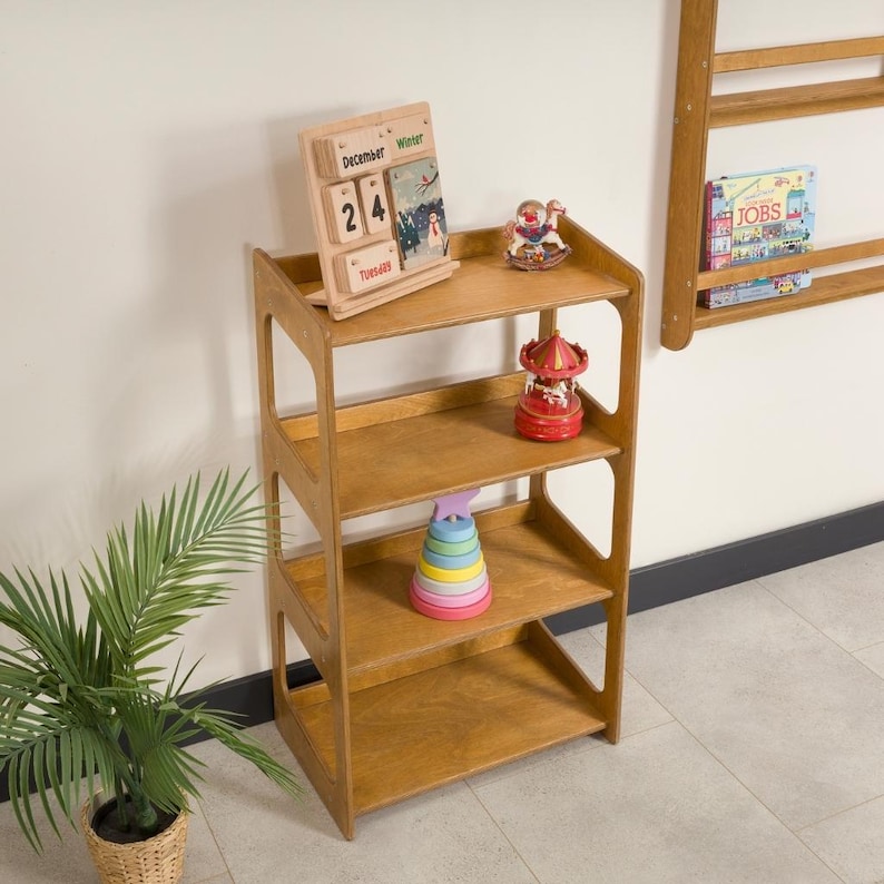 May include: A wooden, four-tiered shelf unit with open sides. The shelves display a wooden calendar, a red carousel toy, and a stack of colorful rings. A small potted plant is in the foreground. A wall-mounted shelf with books is visible in the background.
