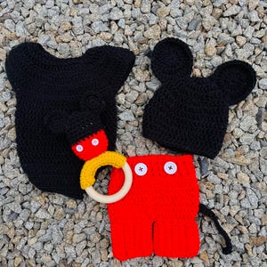 Può includere: Un cappello di Mickey Mouse in crochet nero, pantaloni in crochet rosso con bottoni neri e un maglione con cappuccio in crochet nero con un giocattolo ad anello di legno.