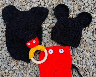 Könnte beinhalten: Eine schwarze gehäkelte Mickey Mouse Mütze, eine rote gehäkelte Hose mit schwarzen Knöpfen und ein schwarzer gehäkelter Kapuzenpullover mit einem Holzringspielzeug.