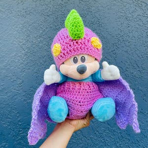 May include: A crocheted purple dinosaur costume with a green spiky head, yellow accents, and blue and white striped sleeves.