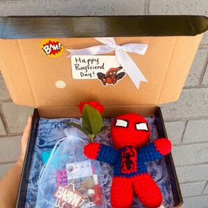 May include: A brown cardboard box with a white ribbon and a "Happy Boyfriend Day!" card featuring a cartoon Spiderman. The box is open and contains a red and blue crocheted Spiderman doll, a red rose, a small bag with a "Marvel" sticker, and a small pink card.